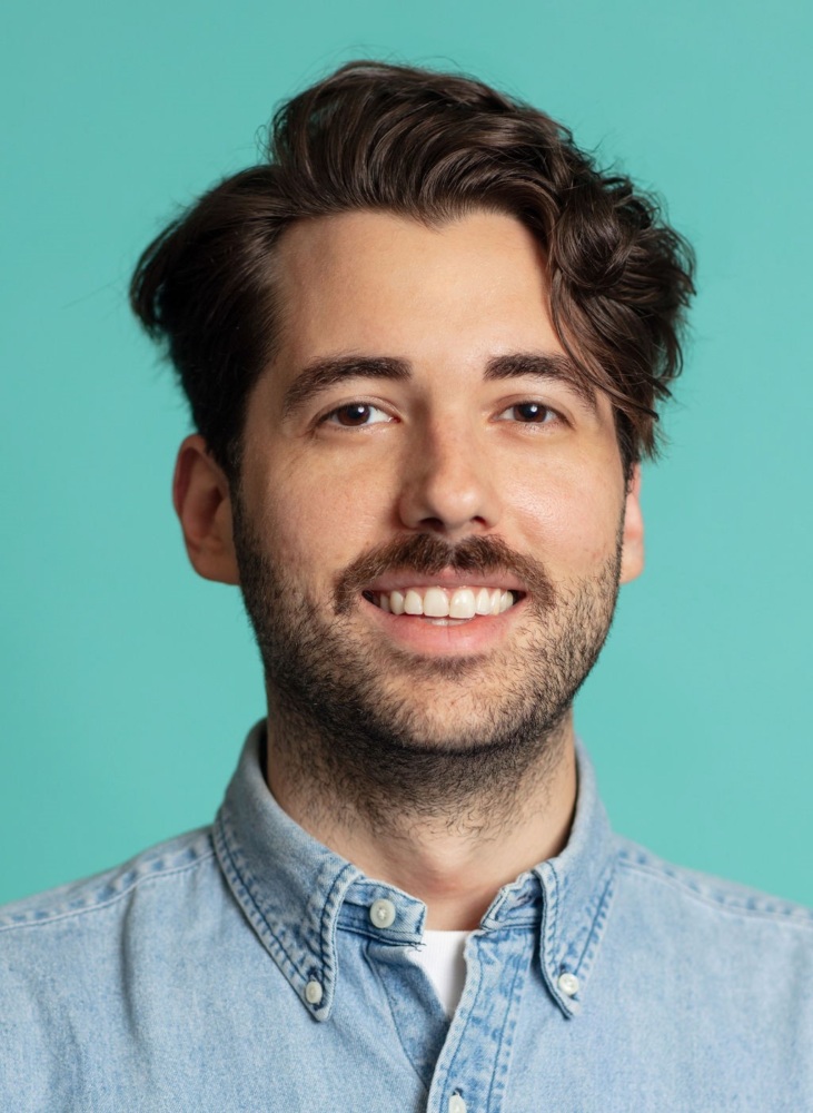 A man with wavy dark hair and a beard is smiling against a turquoise background, wearing a light blue denim shirt.